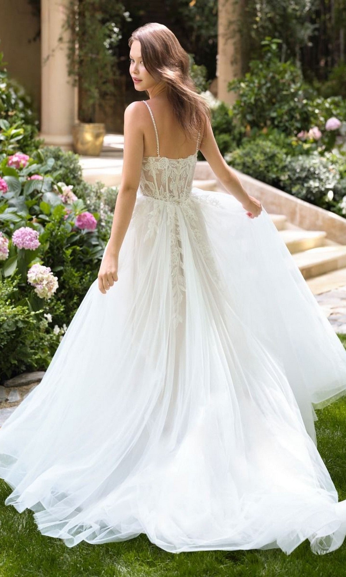 A woman wearing the Andrea and Leo WL087 long wedding dress with a lace back stands in a garden of blooming flowers, her white A-line gown adorned with delicate lace appliques.